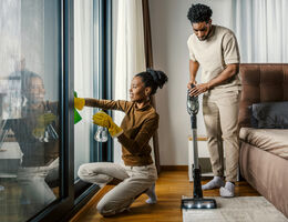In a bedroom, a man vacuums a rug as a woman cleans the window.