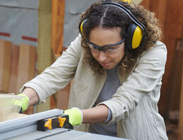 A woman wearing ear protection uses a table saw.