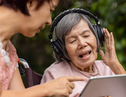 A wheelchair-using older woman is wearing headphones and looking at a tablet held by a younger woman.