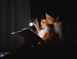 In a dark room, a mom reads to a small boy by flashlight.