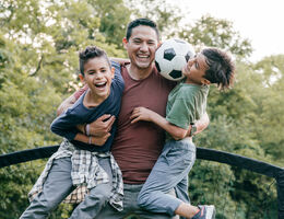 A man holds two laughing boys in front of a soccer goal, squeezing a soccer ball between them.