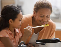 A girl holds up a wooden spoon of vegetable soup for a smiling older woman.