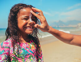 A child grins as an adult applies sunscreen to her nose.