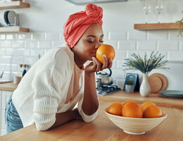 A blissful woman leans on a counter, sniffing an orange from a bowl of them.