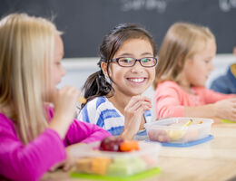 In a row of young students, a girl looks up from her snack and smiles.