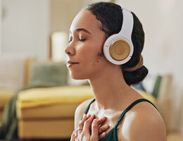 A woman wearing headphones with her eyes closed holds her hands to her chest.
