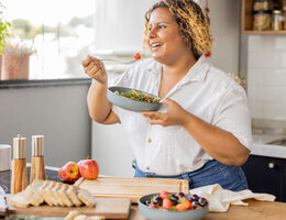 A woman eats a bowl of salad while standing at a kitchen counter.