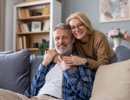 An older woman hugs a gray-haired man on a couch.